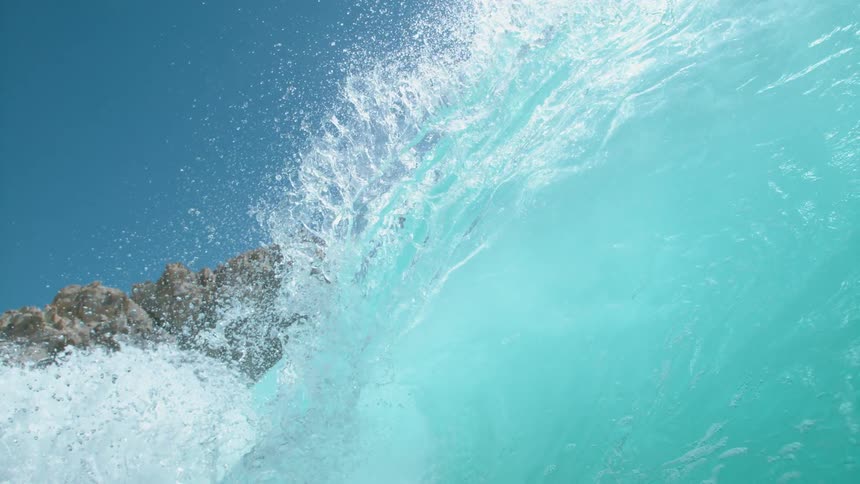 Big blue wave splashes and rolls overhead near rock formations under a clear blue sky