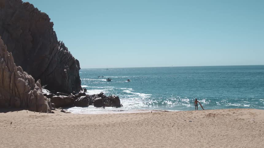 Man drags a surfboard across the beach near rock formations