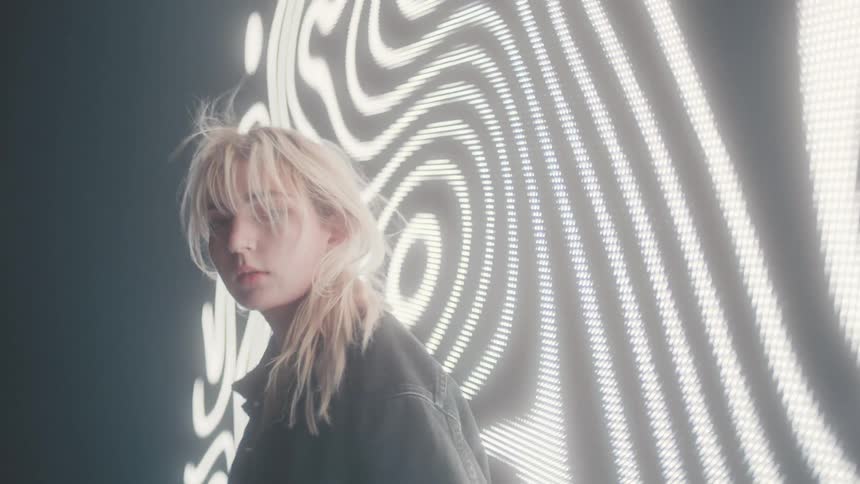 A young blonde woman modeling in front of an electronic sign billboard on the wall of a smoky dark room