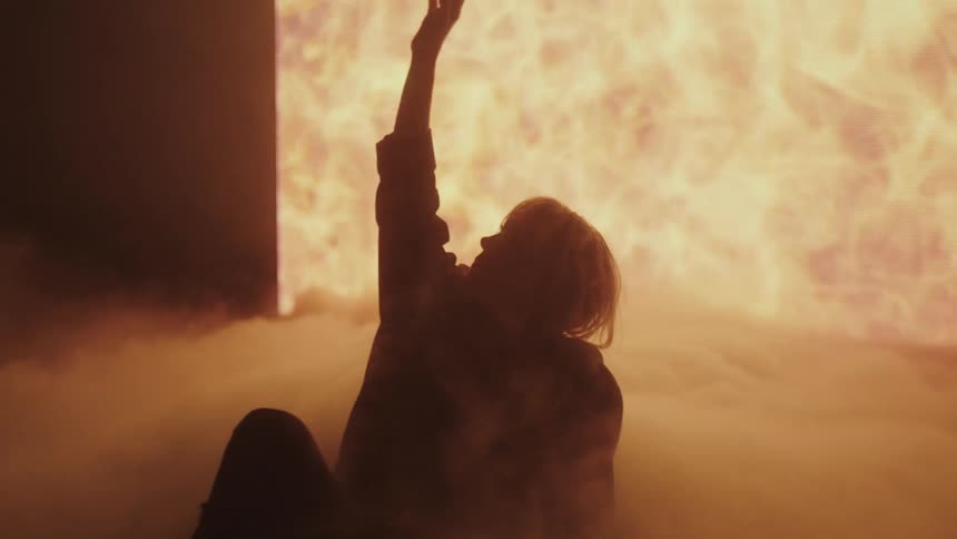 A woman sitting on a stage with smoke with a big screen in the background