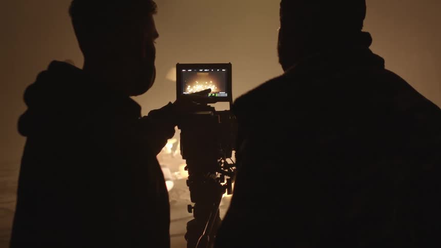 Two men wearing face masks consult over image shown on screen taken from set full of lamps