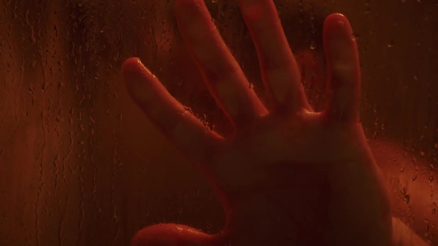 A man touches the glass wall of his shower while showering under a red tinted light