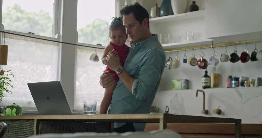 Man holding toddler in arms types on laptop while standing in kitchen