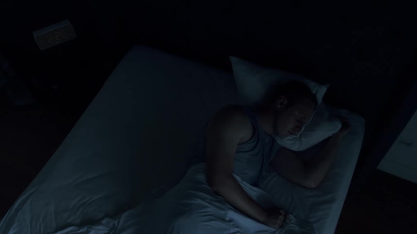 A man asleep on his side in bed with hand under a folded pillow wakes and looks straight ahead