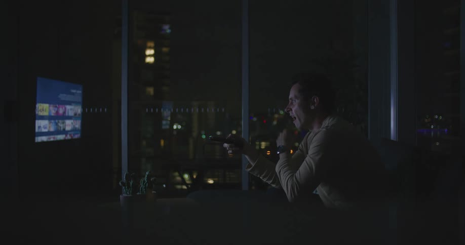 A man yawns while channel surfing on a television in a dark living room