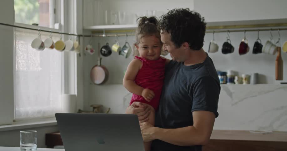 A man in a kitchen holding a baby girl while looking at a laptop on a table