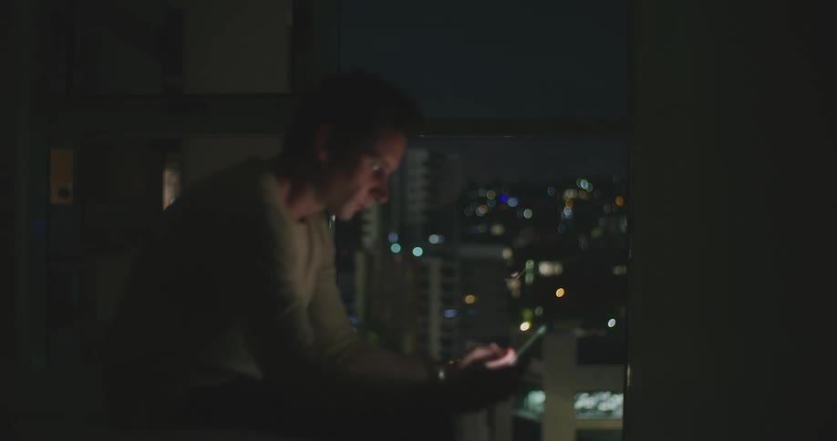 A man lying on a bed in a dark room sits up and browses his smartphone