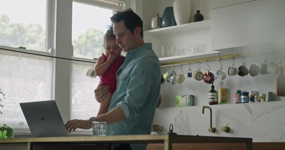 A man types on a laptop with one hand while holding his daughter