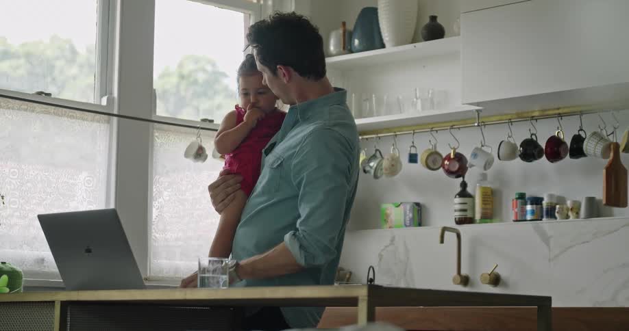 A man in a kitchen holding a baby girl showing her a laptop on a table