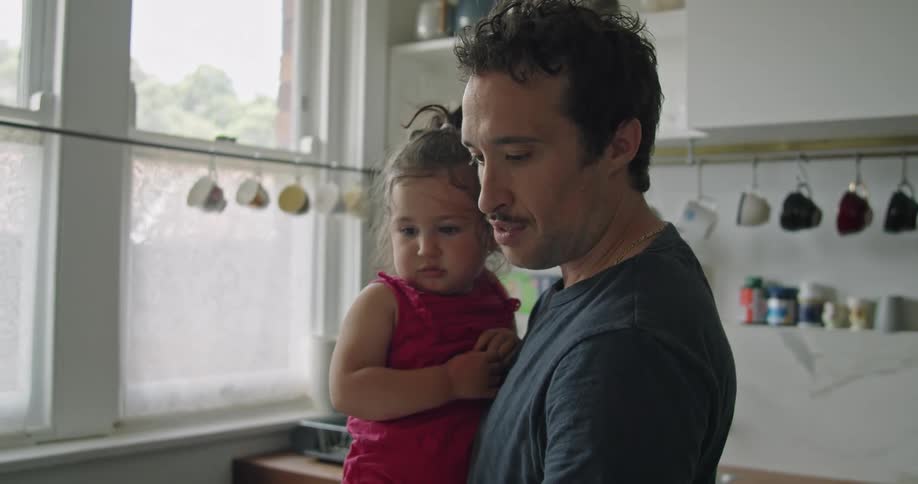 A father holding his daughter with one hand while he uses his other hand to scroll on a laptop on a counter in a kitchen at home during the day
