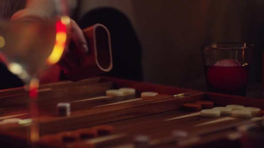 A woman shakes a cup of dice while playing backgammon at home with alcoholic drinks