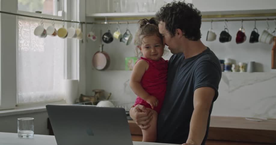 A man holds his daughter and kisses her cheek as they chat to someone during a video call on a laptop
