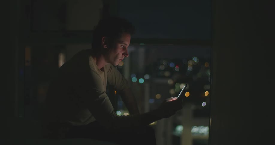 Man with grey sweatshirt uses smartphone in the dark room with illuminated city buildings in the background