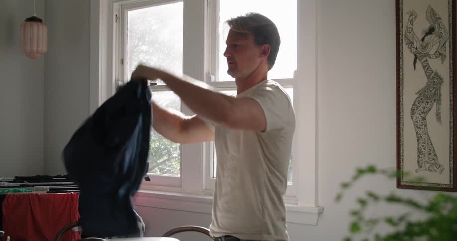 Man with white t shirt and jeans does the ironing and wears the blue shirt