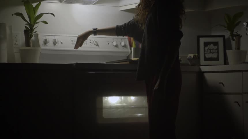 A woman adjusts the settings on a stove in a dimly lit kitchen