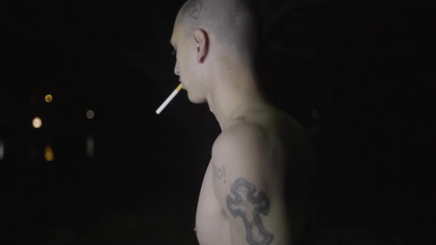 A shirtless man stands looking around with a cigarette in his mouth
