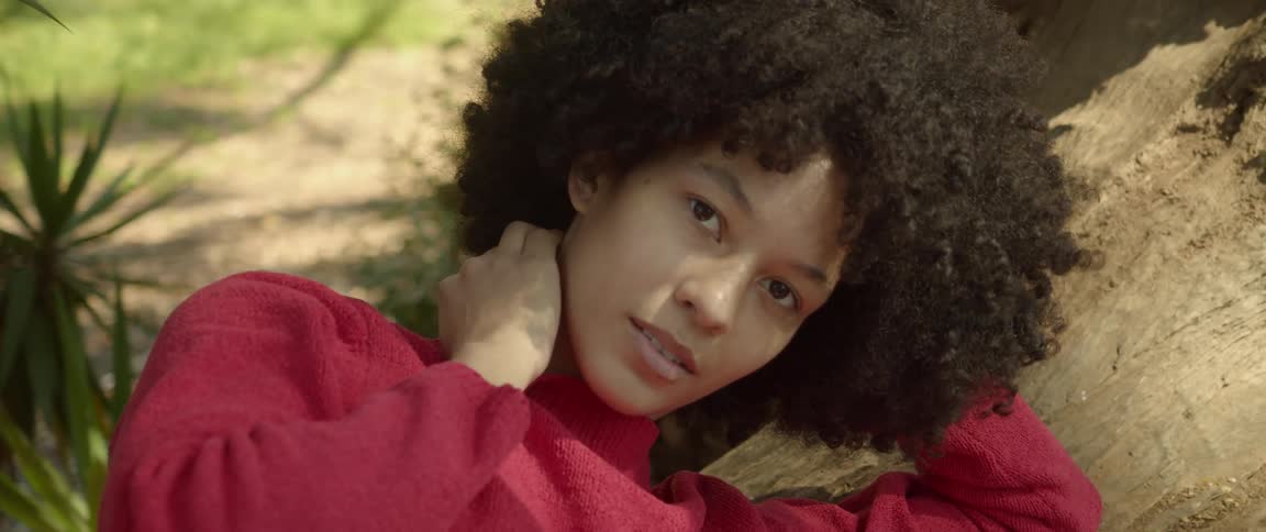 A young woman runs her hand along her neck as gazes into the camera while lying on the trunk of a tree