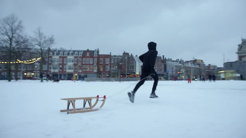 A teenager pulling a sled while running through a snowy park in a small town