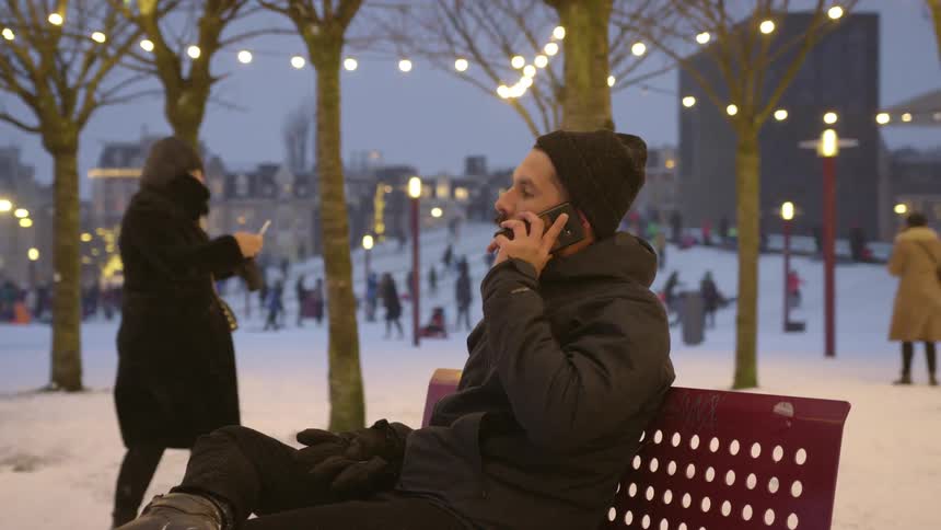 Man sitting in a bench on a park covered in snow takes a call on his phone