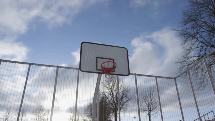 A basketball court with a fence and trees in the background with the sky and clouds above