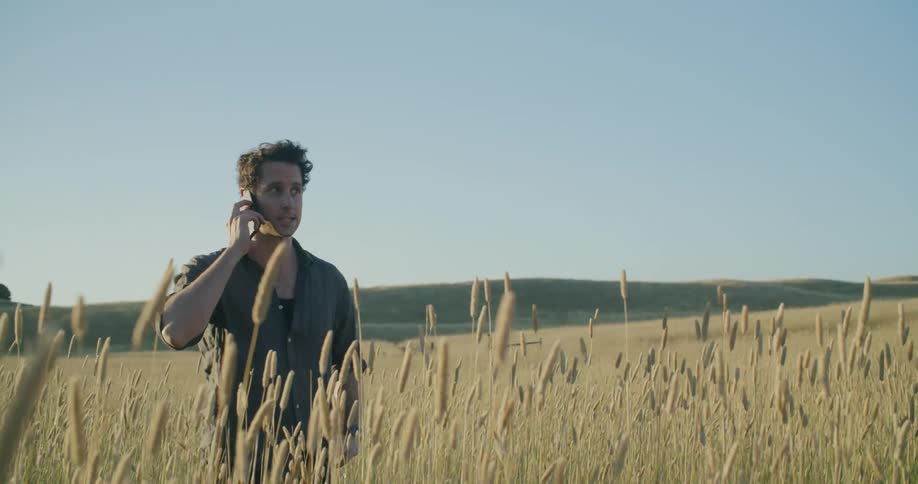 Man stands in field taking on phone on a bright sunny day with hills in the background