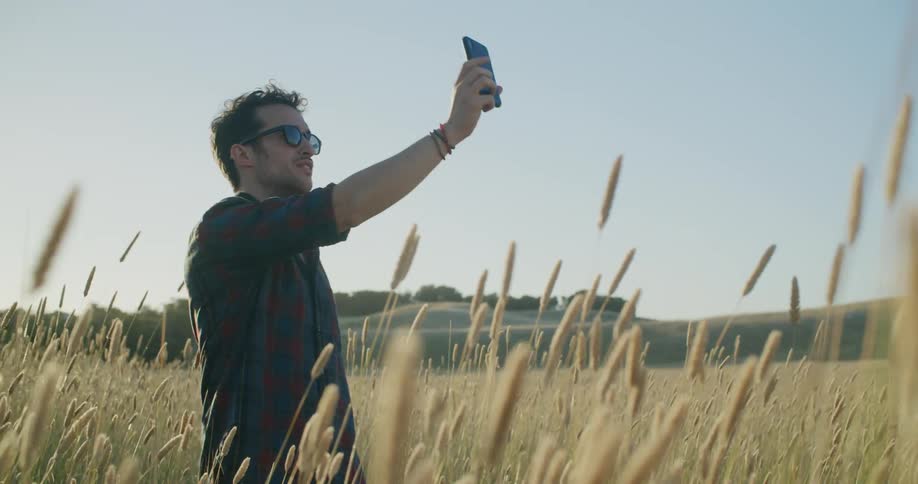 A man with sunglasses and a smart phone in a wheatfield with trees the sky and clouds in the background