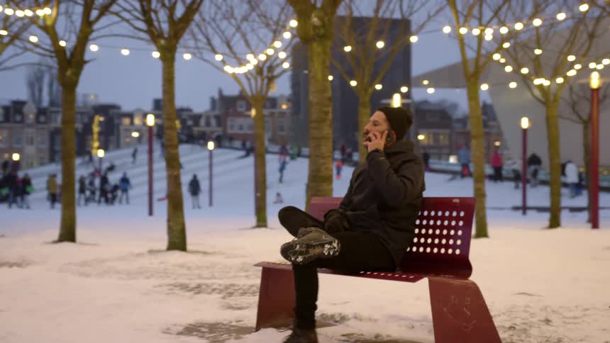 Man sitting on a bench in the snow covered park in the city answers a call on his phone