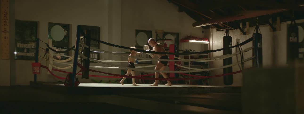 A strong muscular man sparring with a young boy on a boxing ring inside a gym