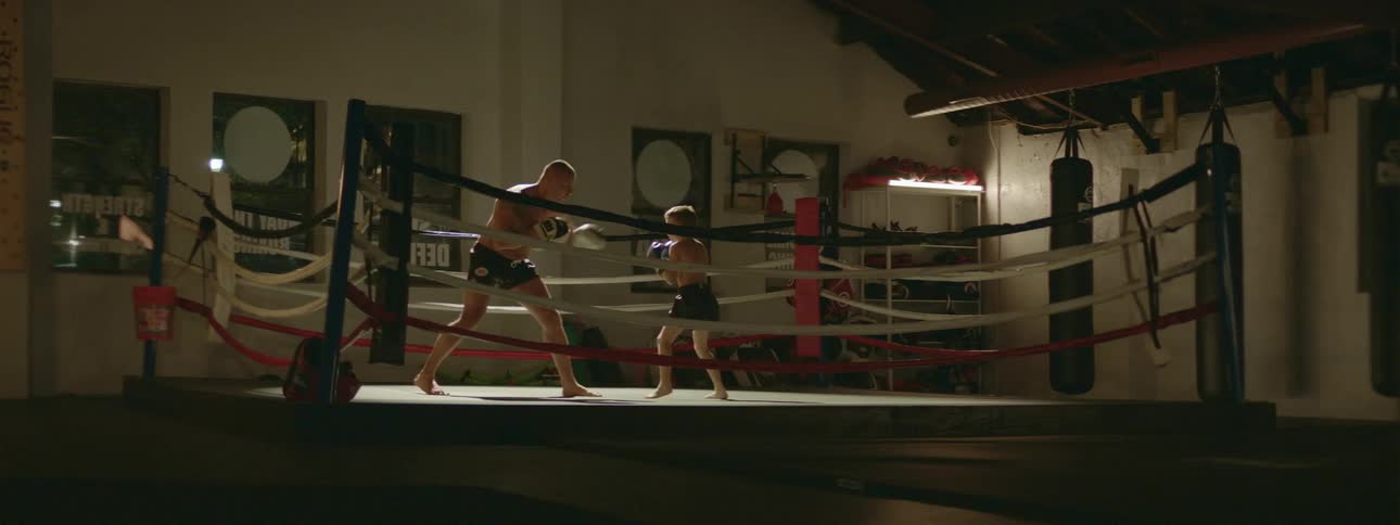Man teaches child how to box in a boxing ring with posters and a heavy bag in the background