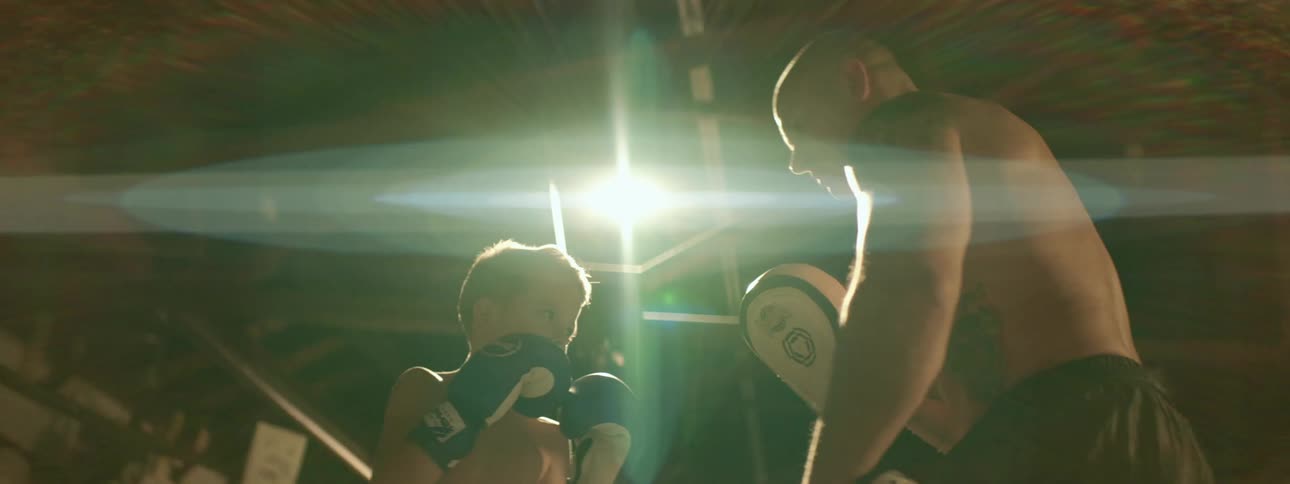 A young boy wearing boxing gloves trains with a strong muscular shirtless man inside a gym