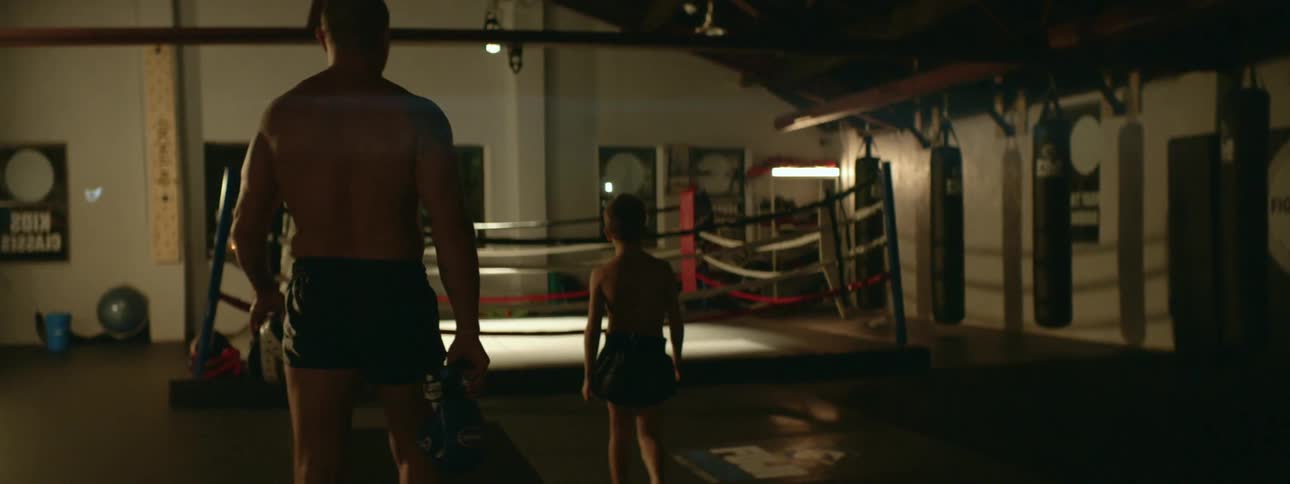 A man carrying boxing gloves and pads walks towards a boxing ring with a boy in a gym