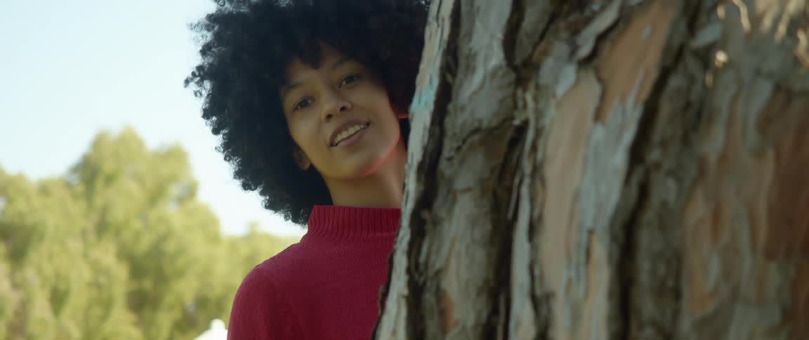 A woman staring and smiling while peeking from behind a tree with trees the sky and clouds in the background
