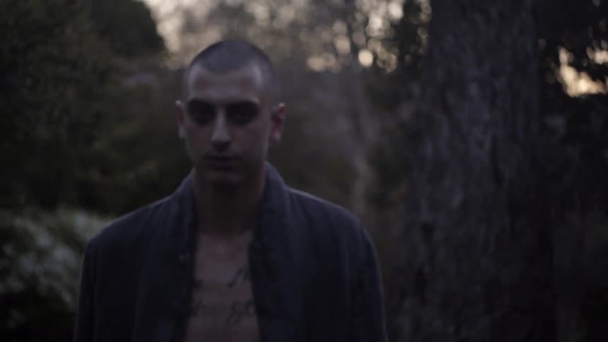 A man with his shirt open walks towards the camera in the evening