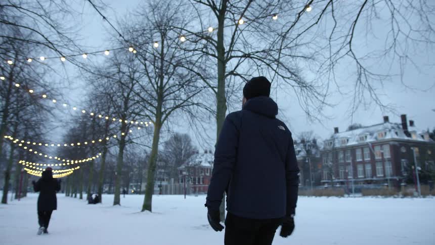 A man walking under decorative lights hanging from trees on the snow covered boulevard in the city