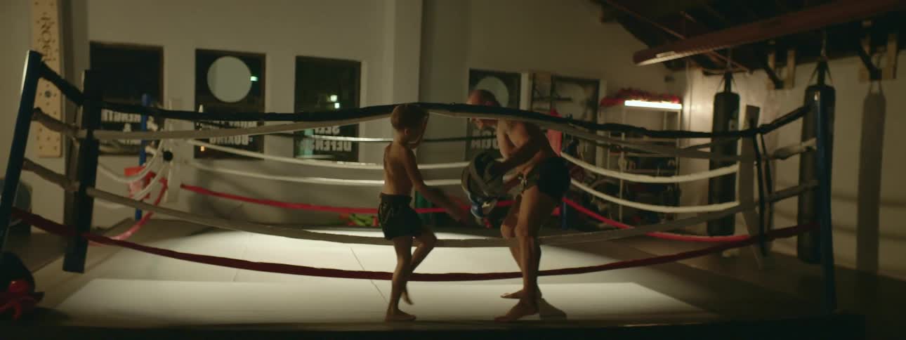 A young boy and a man walk towards a boxing ring and they both hold the ropes for one another as they climb in to the ring