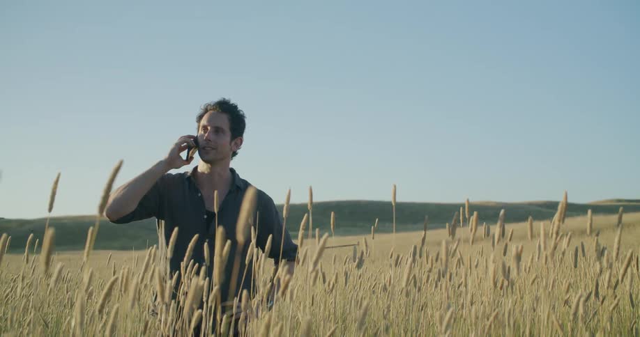 Man stands in field talking on the phone with hills in the background on sunny day with a bright blue sky