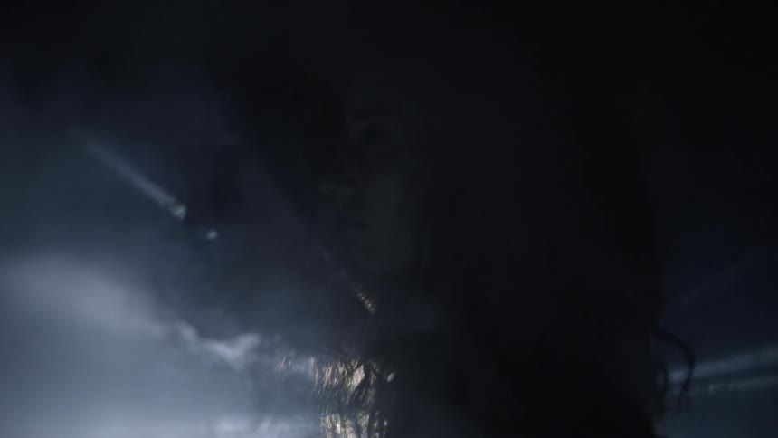 Woman stands in darkness as lights flash and smoke moves towards her