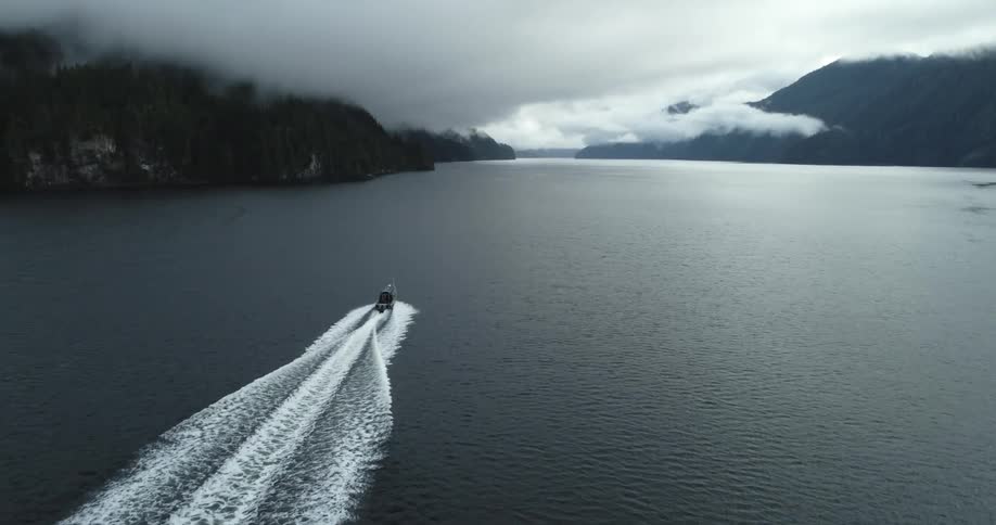 Boat cuts through lake toward forest covered mountains obscured by low lying clouds