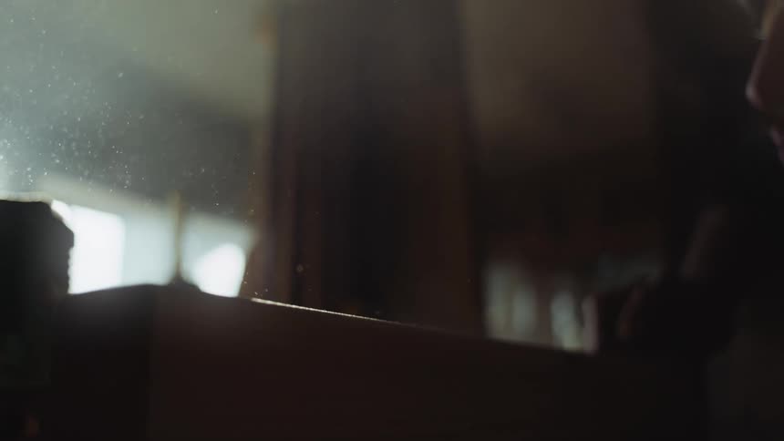 Man sands wood down creating dust that shows in the sunlight coming from the window