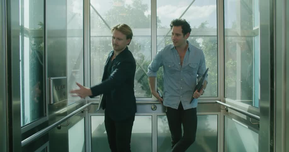 A man gets onto a glass elevator where his coworker is standing with a laptop and smartphone
