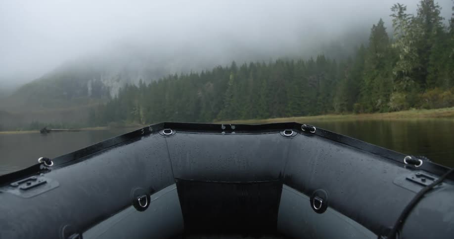 A black boat floating on a lake surrounded by forest trees on a cloudy day