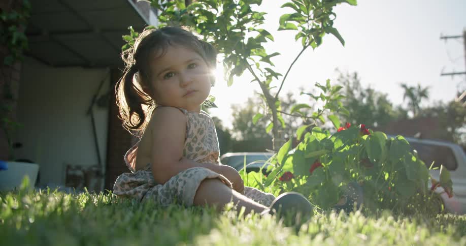 Little girl sits on grass in front of plants with cars and a house in the background