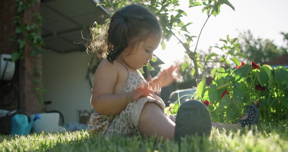 A baby girl sitting on grass with plants a house the sky and clouds in the background