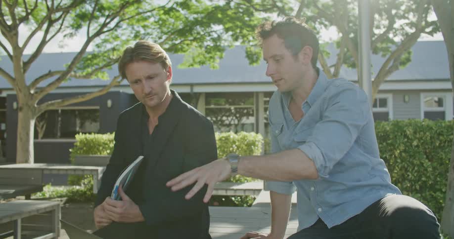 Two men sitting on a table looking at a laptop with trees a building the sky and clouds in the background