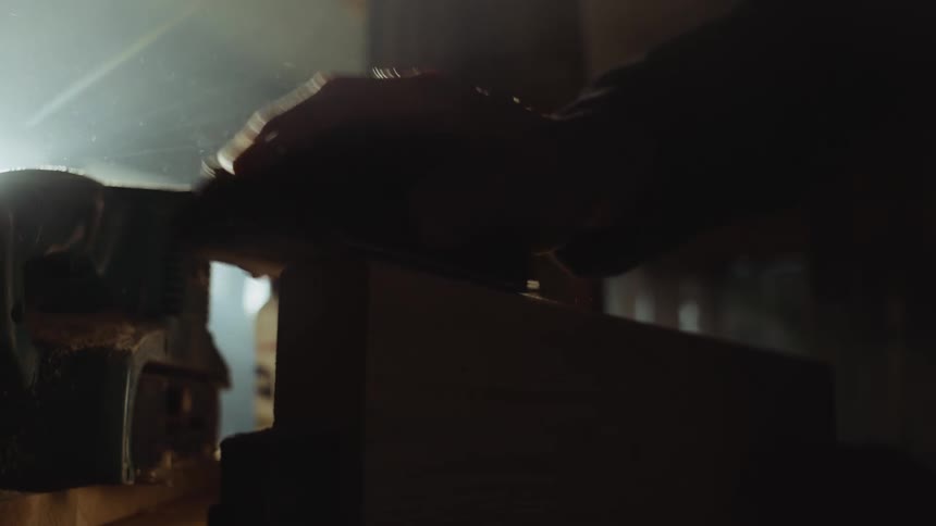 Man shaves wooden block with hand tool in shadowy workshop spreading dust into air