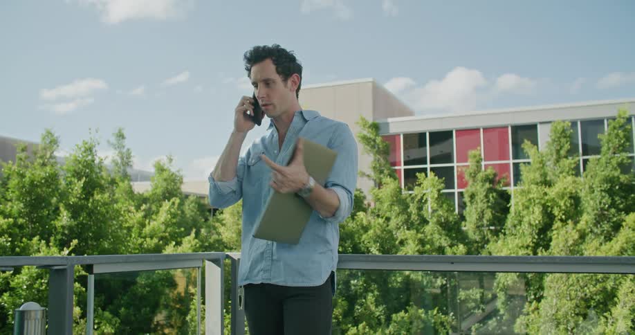 Man stands outside leaning on railing holding a laptop and talking on a cell phone with a building and trees in the background on a sunny day with fluffy clouds