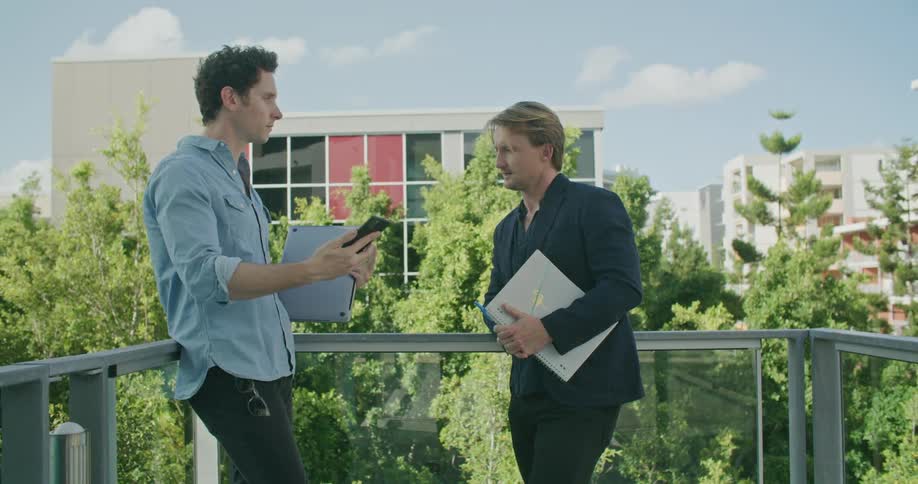 Two man reclined on the terrace of a building talking and looking at a phone