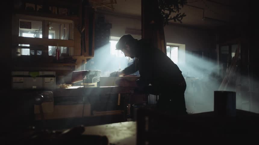 A man sands a wooden board in a dimly lit workshop during the day