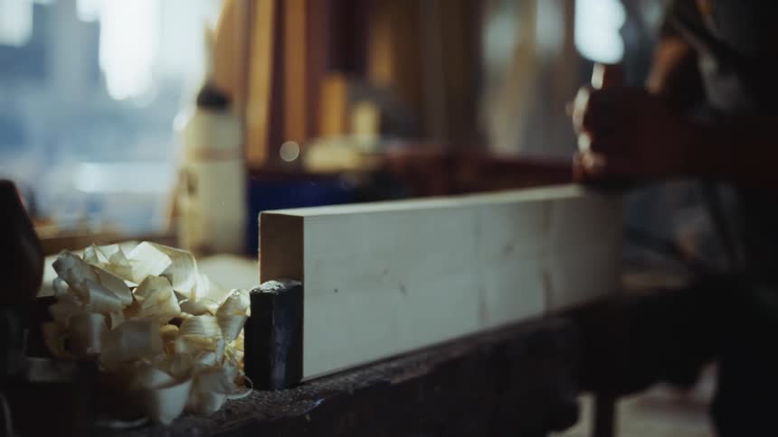 Man works on shaving down a piece of wood in front of some windows