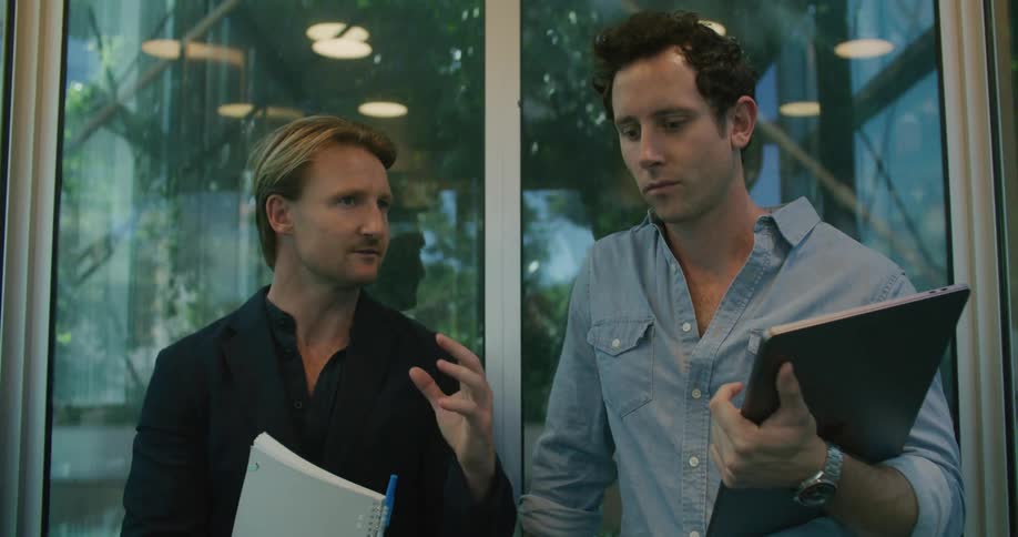 Two men riding an elevator with one holding a notebook and pen and the other holding a laptop